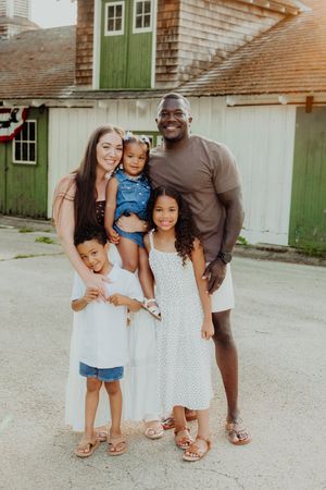 A happy interracial family of five poses outdoors in front of a green barn, smiling warmly in summer attire.