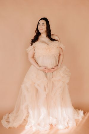 Elegant maternity portrait in flowing peach tulle gown against peachy backdrop with soft studio lighting.