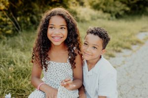 Two smiling children with curly hair pose together outdoors on a sunny day, sitting on a gravel path near green trees.