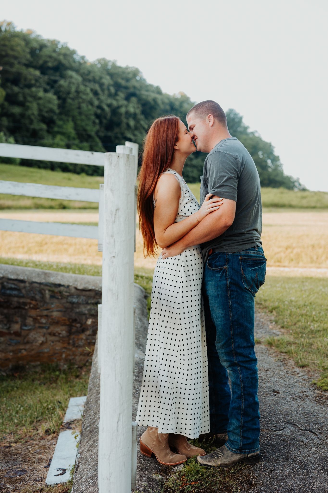 Couple sharing a romantic kiss beside a white fence in a rural countryside setting, woman in polka dot dress.