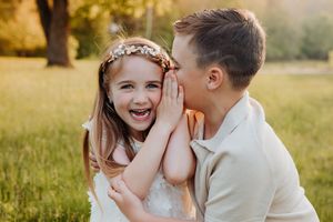 A boy whispers a secret to a laughing girl wearing a floral crown in a sunny green meadow.