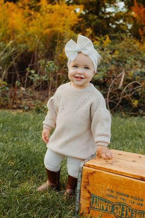 Smiling toddler girl in cream knit sweater and white bow headband standing by vintage wooden crate outdoors in fall.