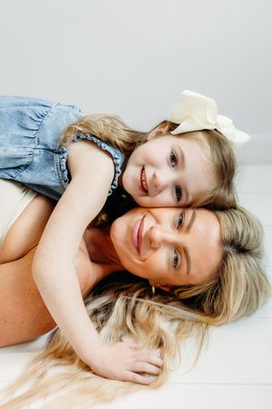 A smiling mother lying down with her young blonde daughter in a denim dress hugging her from behind.