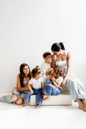 Two women and three children sit together on a white couch, dressed in jeans and white tops, smiling warmly.