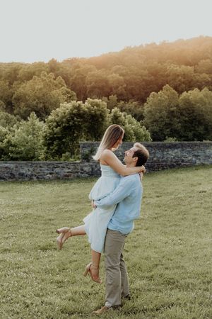 A couple shares a romantic moment as a man lifts a woman in a blue dress on a green lawn at sunset.