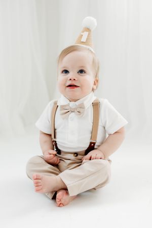 A baby in a white shirt and beige suspenders sits on a white backdrop in a series of poses.