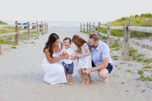 Family of four enjoying a sunny beach day on a sandy path with wooden fences, parents smiling at laughing children.