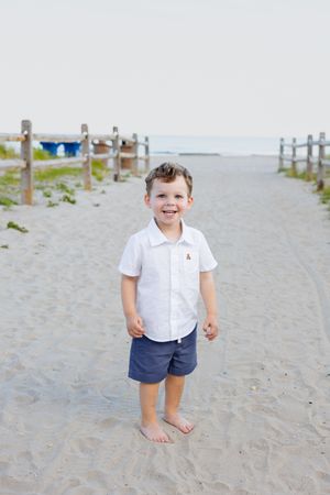 A smiling young boy in a white shirt and blue shorts stands barefoot on a sandy beach path with wooden fencing.
