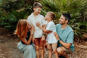 A happy family of four laughs together outdoors surrounded by lush tropical greenery on a sunny day.