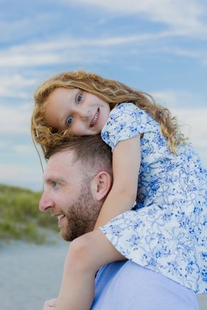 A bearded father giving his curly-haired blue-eyed daughter a piggyback ride on a sunny beach day.