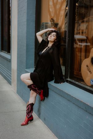 Woman in black dress and red cowboy boots posing against a blue brick wall near a guitar shop window.