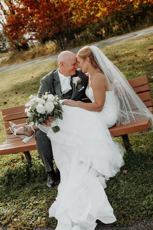 Bride and groom share a romantic moment on a park bench surrounded by vibrant fall foliage, bride holding white bouquet.