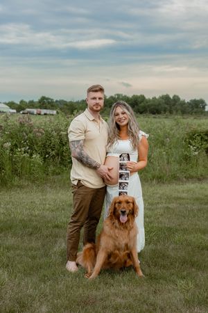 Couple holding ultrasound photos with golden retriever dog in green field, pregnancy announcement photo session outdoors.