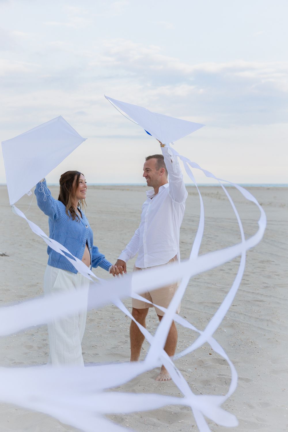 Couple holding a white kite with flowing ribbons on a sandy beach, smiling at each other on a cloudy day.