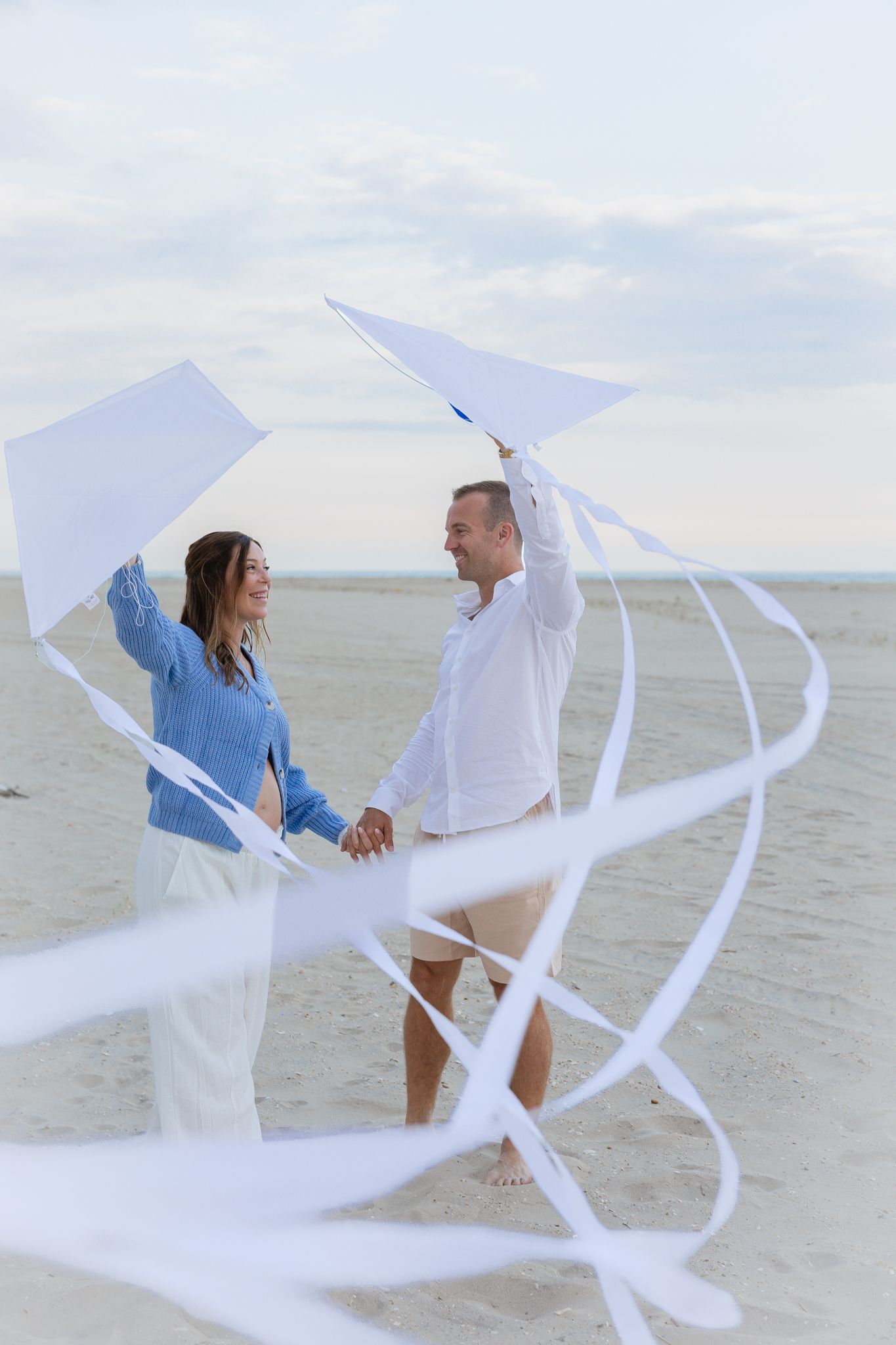 Couple holding a white kite with flowing ribbons on a sandy beach, smiling at each other on a cloudy day.