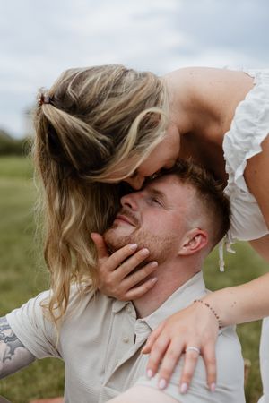 A woman in a white dress kisses a man on the forehead while cradling his face outdoors.