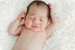 Peaceful newborn baby sleeping on a soft white fluffy blanket with tiny hands raised beside head.