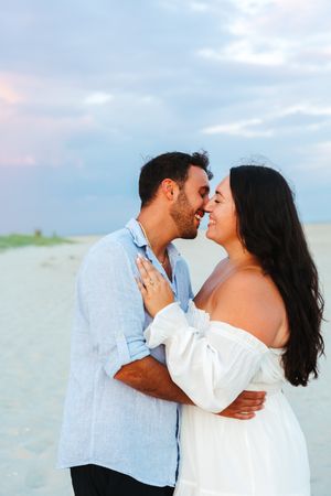 Couple sharing a romantic almost-kiss on a sandy beach at sunset, woman in white off-shoulder dress, man in light blue shirt.