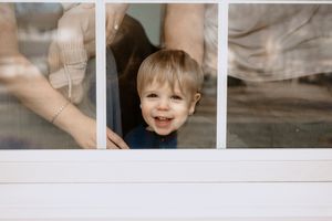 Smiling toddler boy peering through a window with adults standing behind him on a bright day.