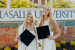 Two smiling female graduates in white gowns holding caps in front of La Salle University brick sign.