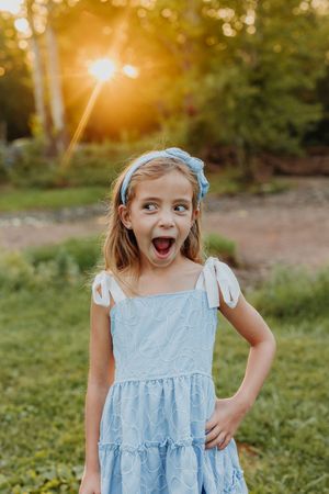 A young girl in a blue dress with white bow shoulders and blue headband laughs outdoors at golden sunset.