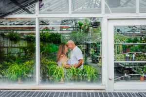 Couple sharing a romantic moment inside a lush green greenhouse, surrounded by tropical plants visible through glass walls.