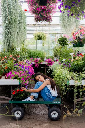 Young girl sitting on a garden cart surrounded by colorful flowers and plants in a lush greenhouse.
