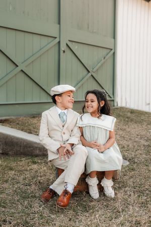 Two dressed-up children sit together outside a barn, boy in tan suit and cap, girl in green gingham dress.