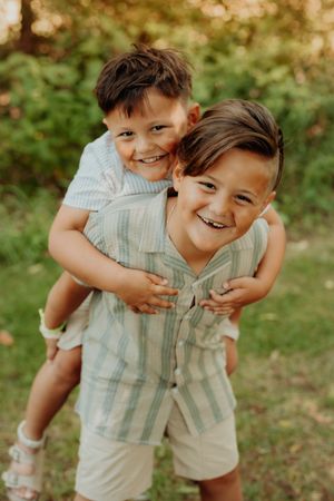 Two playful kids in matching plaid shirts and light shorts share joyful hugs on a sunny day in a grassy backyard.
