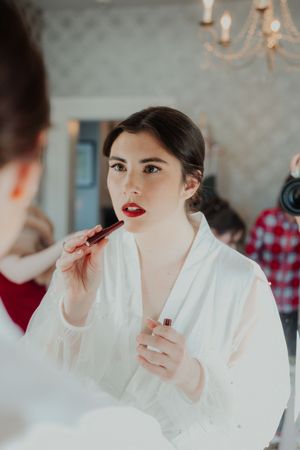 Makeup artist applies bright red lipstick on a person wearing a white robe during beauty preparation.
