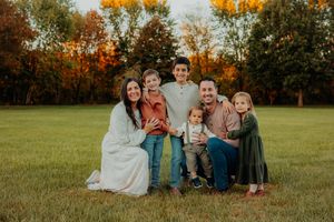 Family portrait in a grassy field with autumn trees displaying vibrant orange and yellow foliage in the background.