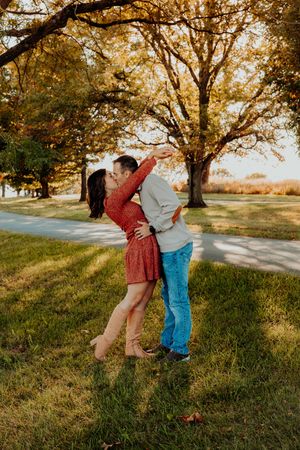 A couple in casual attire embraces and dances together on grass under golden autumn trees at sunset.