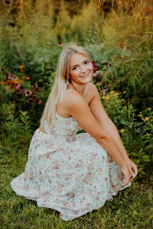 A person in a floral dress sits on grass surrounded by blooming wildflowers in a garden setting during golden hour.