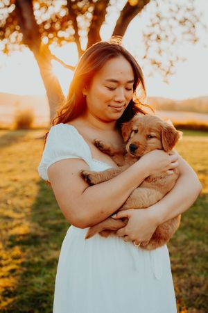 A person in a white dress holds and cuddles with a golden retriever puppy during golden hour sunset outdoors.