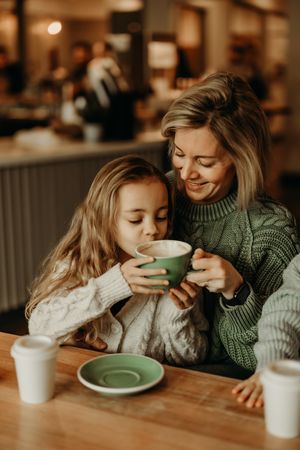 Two people in cozy sweaters share intimate moments while drinking coffee at a wooden table in warm lighting.