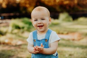 A smiling infant wearing blue denim overalls and a white t-shirt stands outdoors during autumn.