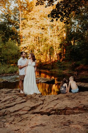 A couple in white outfits sits along a rocky riverbank at sunset with golden light streaming through autumn trees.