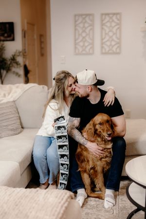 A couple sits on a white couch with their golden retriever while holding ultrasound photos in a home setting.