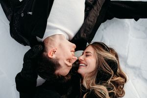 A couple lies on a white surface wearing black clothing while laughing together with their heads touching.