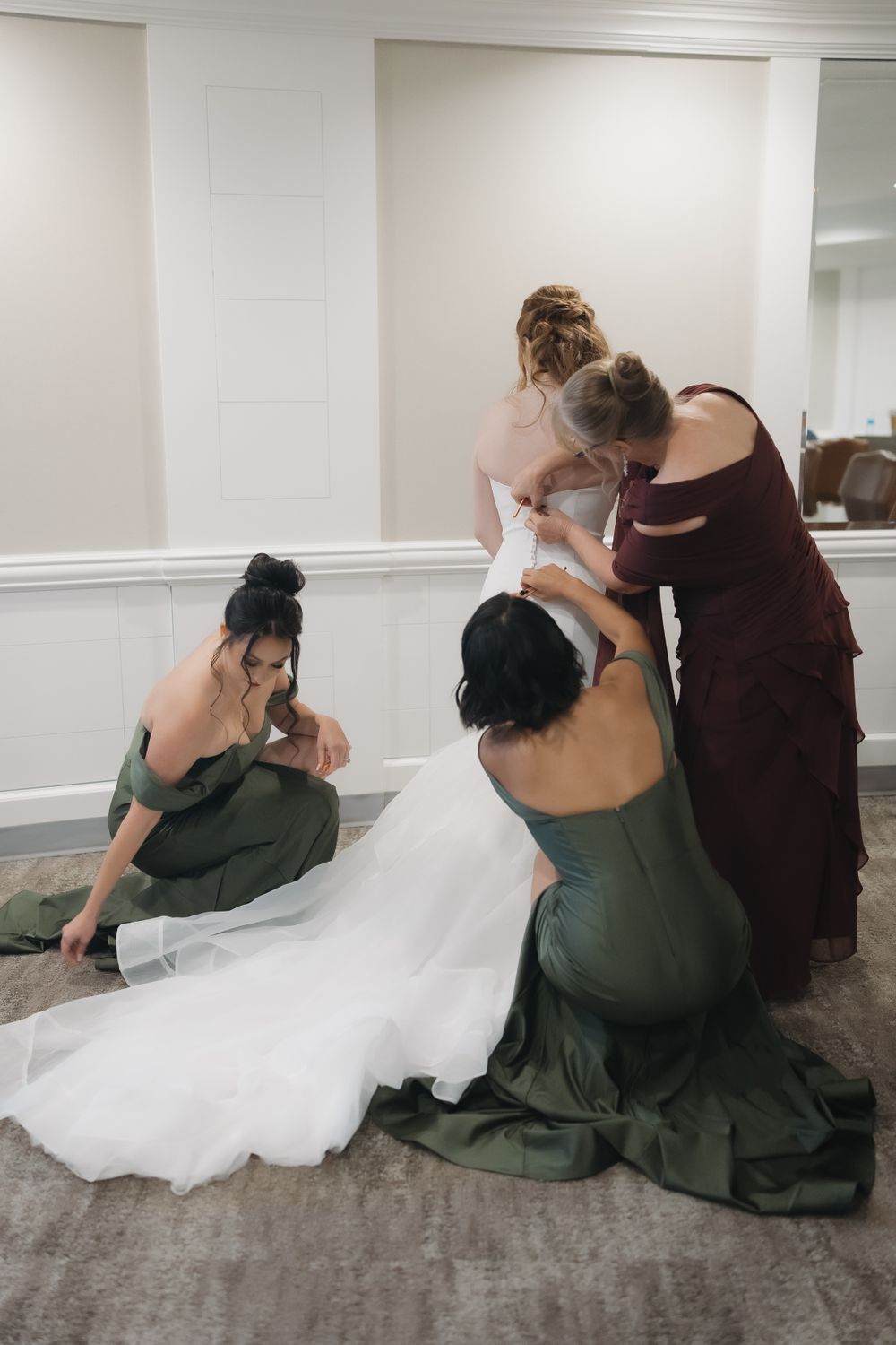 Bridesmaids in dark green dresses help adjust a wedding gown's train on the floor.