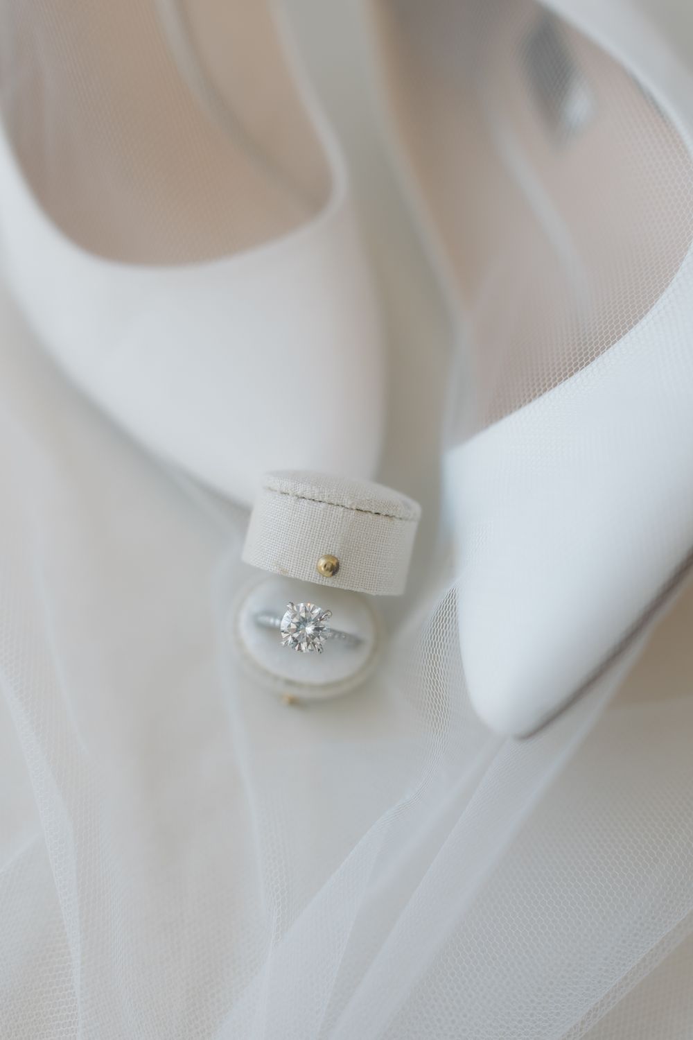 Close-up details of white bridal shoes and veil against light background.
