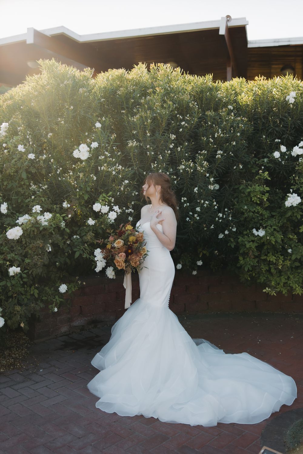 A bride in a flowing white gown poses against a garden backdrop at golden hour.