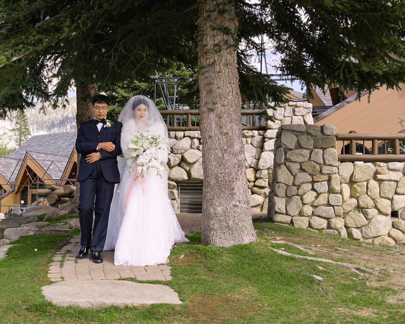 A bride is taken down the path by her father to the wedding ceremony site. Rustic stone wall is in the background.