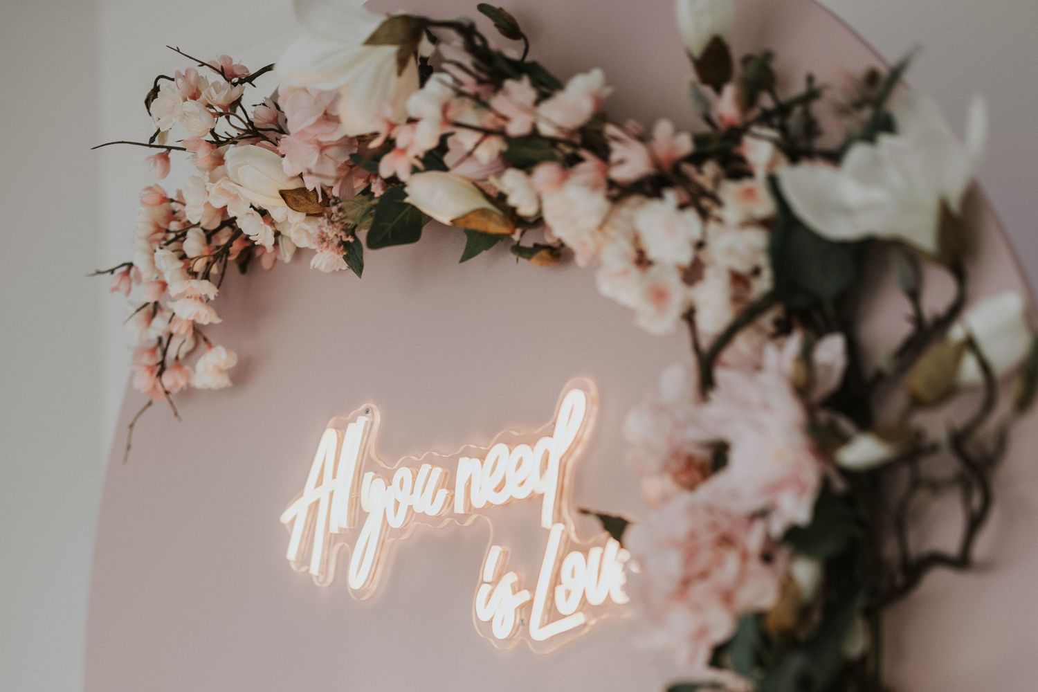 A neon sign reading All you need is Love surrounded by pink cherry blossom flowers on a blush wall.
