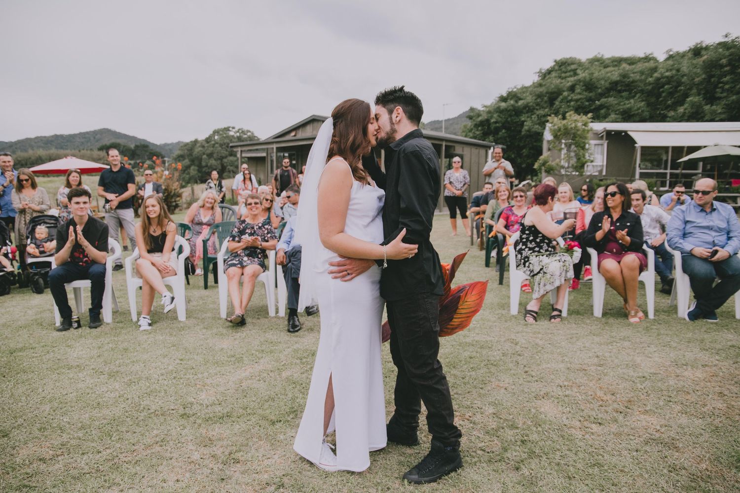 A couple shares their first kiss as newlyweds on a grassy lawn surrounded by wedding guests at an outdoor reception.