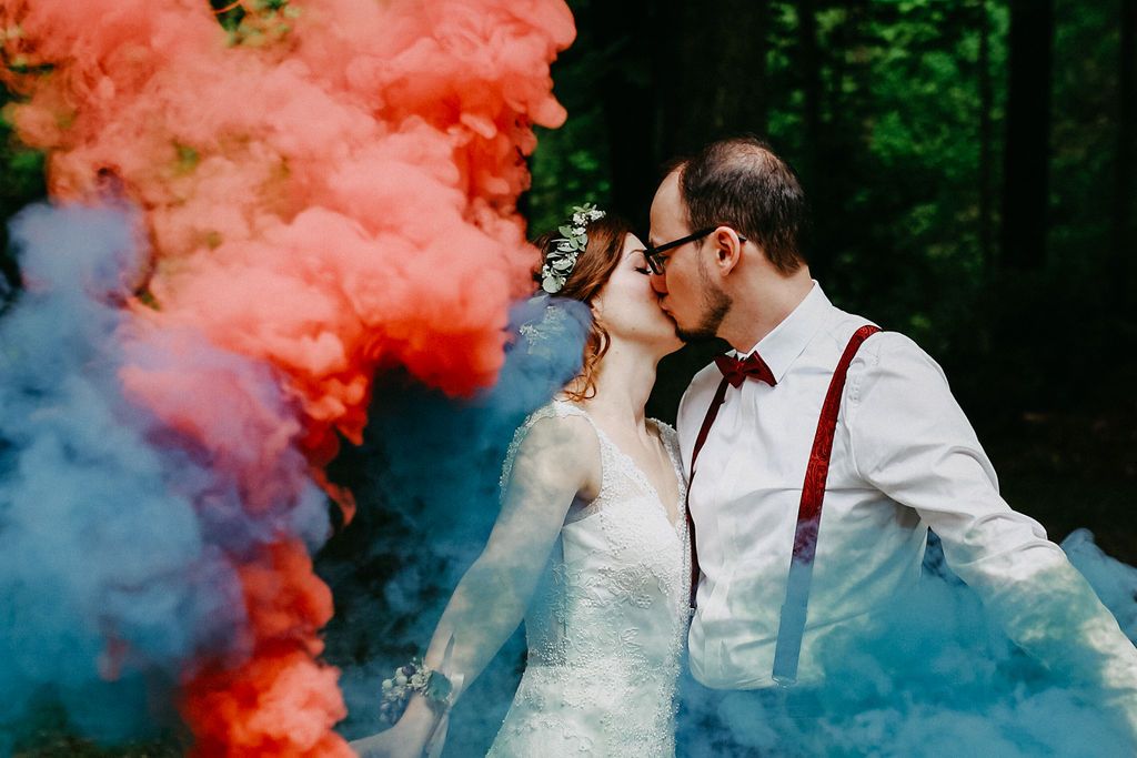 Wedding couple kisses surrounded by dramatic pink and blue smoke bombs in a forest setting.