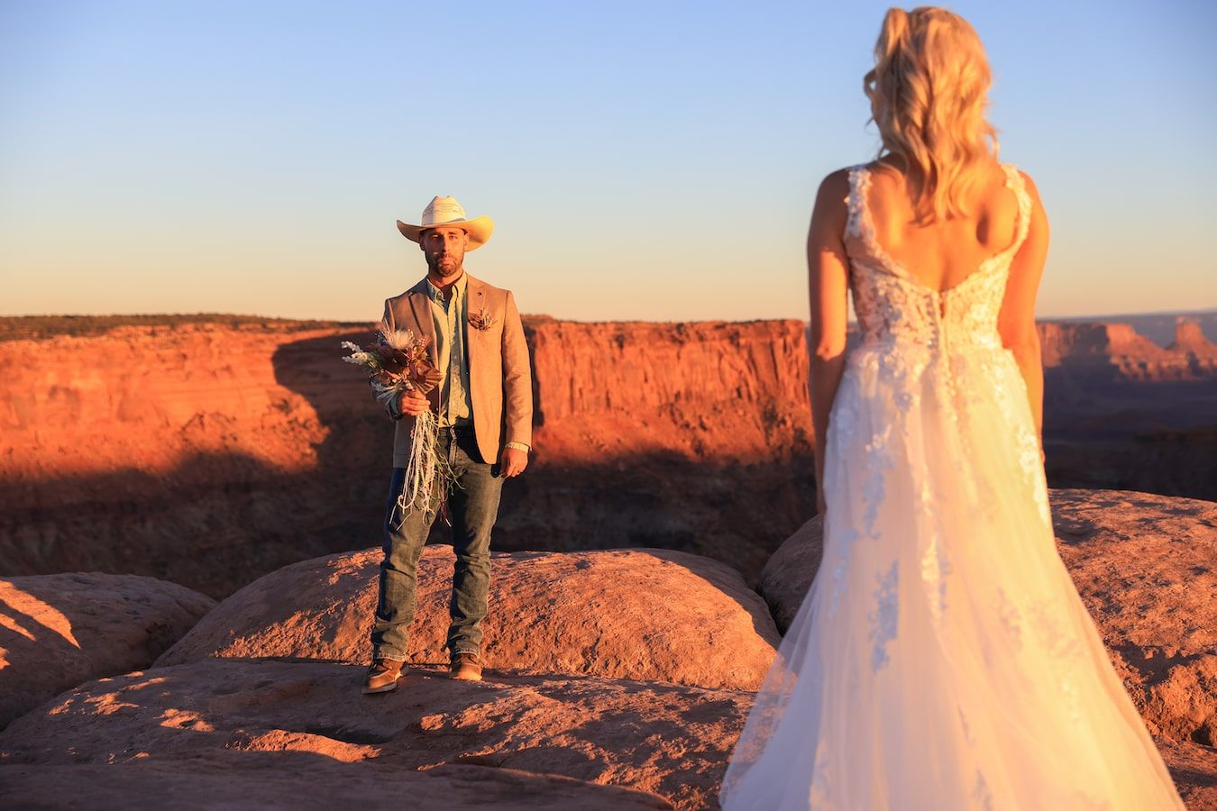 Two people stand on red rock formations during a golden sunset in what appears to be the American Southwest.