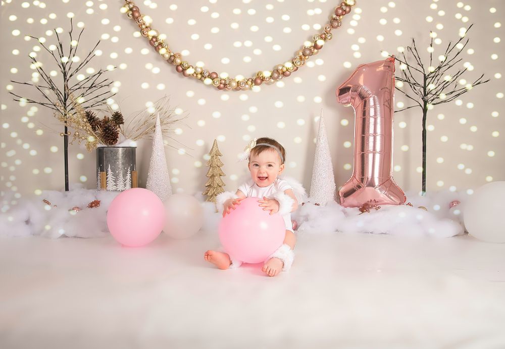 Pink and gold first birthday photo setup with winter trees, balloons, and sparkly lights in the background.