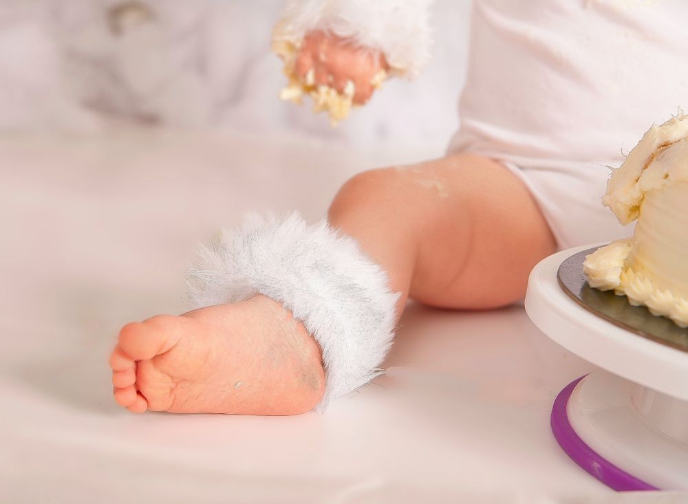 Baby wearing white fluffy anklet with a slice of cake on a plate nearby.