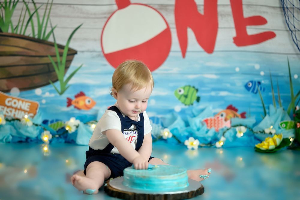 A baby in navy overalls sits with a blue cake against an underwater-themed backdrop with colorful fish decorations.